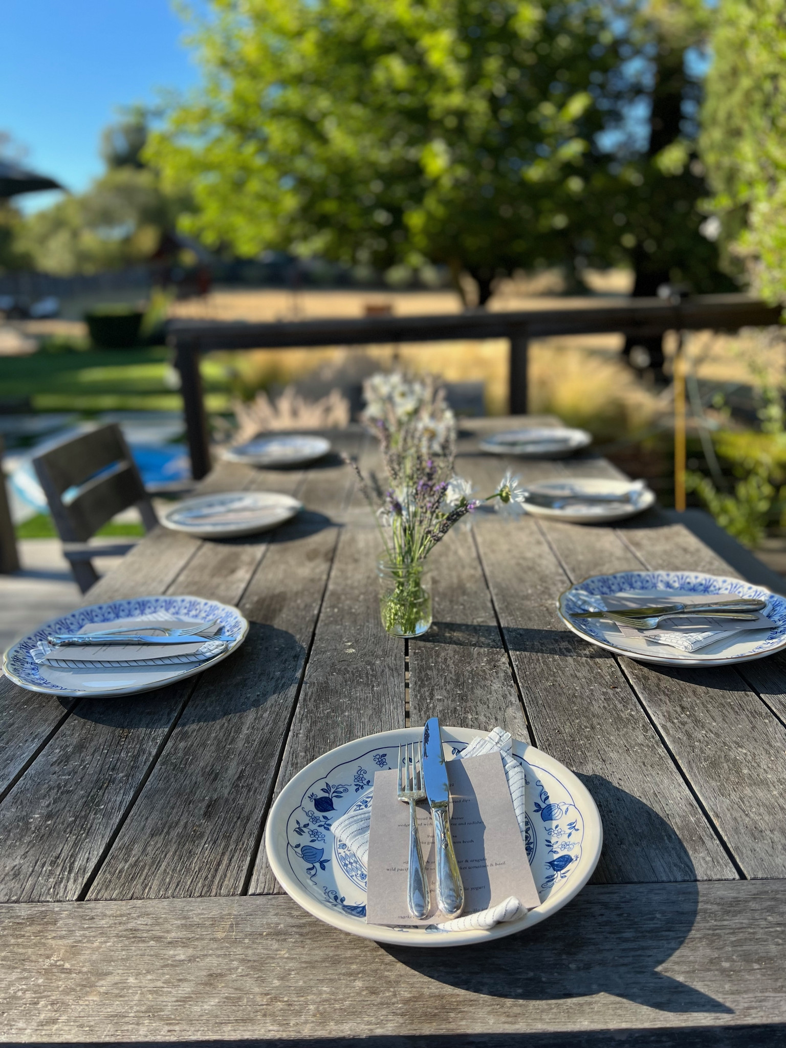 An outdoor table set for dinner with flowers, in a bright green field